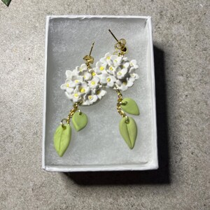K&ouml;nnte beinhalten: Ein Paar wei&szlig;e Blumenohrringe mit gr&uuml;nen Bl&auml;ttern, die an goldenen Ketten h&auml;ngen. Die Ohrringe werden in einer wei&szlig;en Schachtel pr&auml;sentiert.
