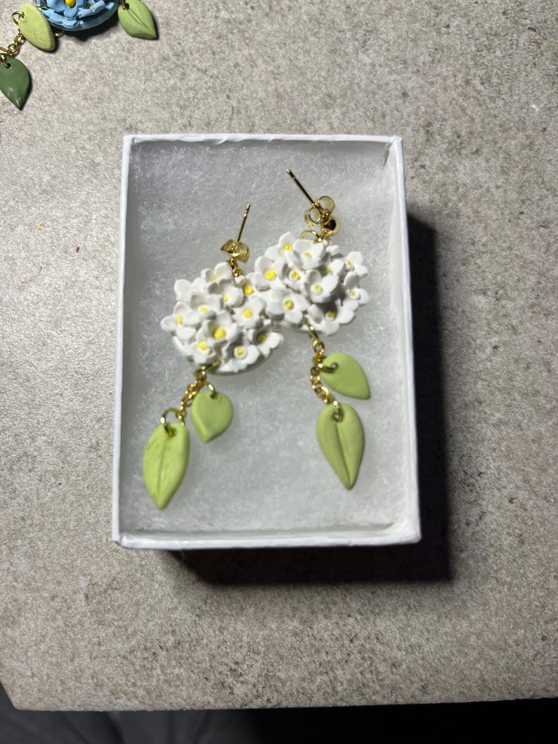 K&ouml;nnte beinhalten: Ein Paar wei&szlig;e Blumenohrringe mit gr&uuml;nen Bl&auml;ttern, die an goldenen Ketten h&auml;ngen. Die Ohrringe werden in einer wei&szlig;en Schachtel pr&auml;sentiert.