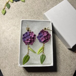 K&ouml;nnte beinhalten: Ein Paar violette und gr&uuml;ne Blumenohrringe mit goldenen Ketten und h&auml;ngenden Bl&auml;ttern. Die Ohrringe werden in einer wei&szlig;en Schachtel pr&auml;sentiert.