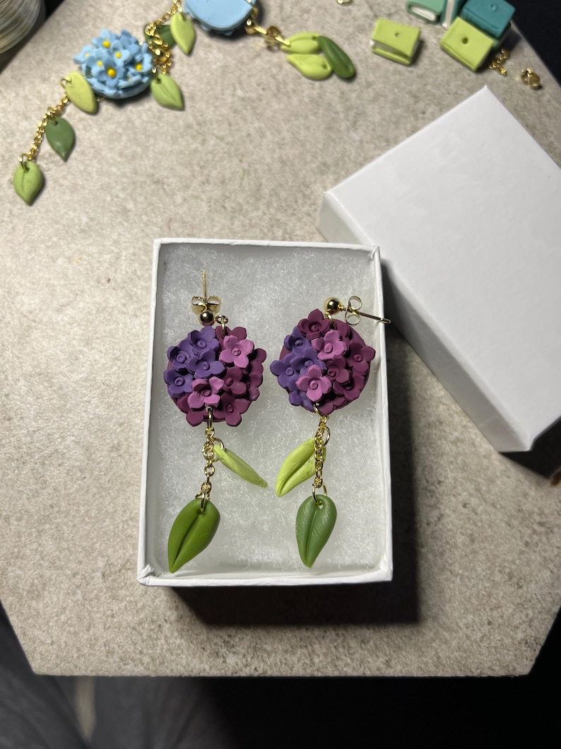 K&ouml;nnte beinhalten: Ein Paar violette und gr&uuml;ne Blumenohrringe mit goldenen Ketten und h&auml;ngenden Bl&auml;ttern. Die Ohrringe werden in einer wei&szlig;en Schachtel pr&auml;sentiert.