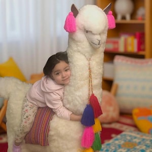May include: A young girl is riding a white stuffed alpaca with pink ear tassels. The alpaca has a colourful tassel trim. The girl is wearing a pink shirt and floral trousers.
