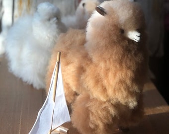 May include: A brown alpaca plush toy with white fur around its face and ears, sitting on a wooden surface with a small white sailboat toy in front of it.