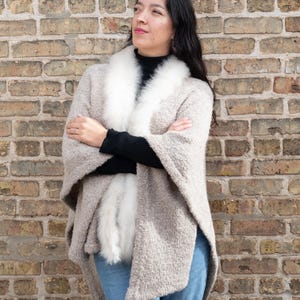 May include: A woman wearing a beige cape with a white fur collar and trim, paired with a black top and blue jeans. The cape has a textured appearance. The background is a brick wall.