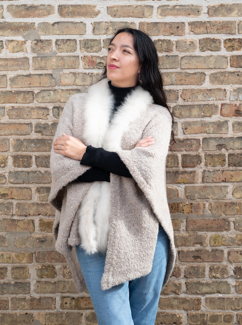 May include: A woman wearing a beige cape with a white fur collar and trim, paired with a black top and blue jeans. The cape has a textured appearance. The background is a brick wall.