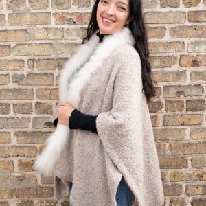 May include: A beige poncho with a white fur collar. The poncho has a textured appearance and is worn over a black top and blue jeans. The background is a brick wall.