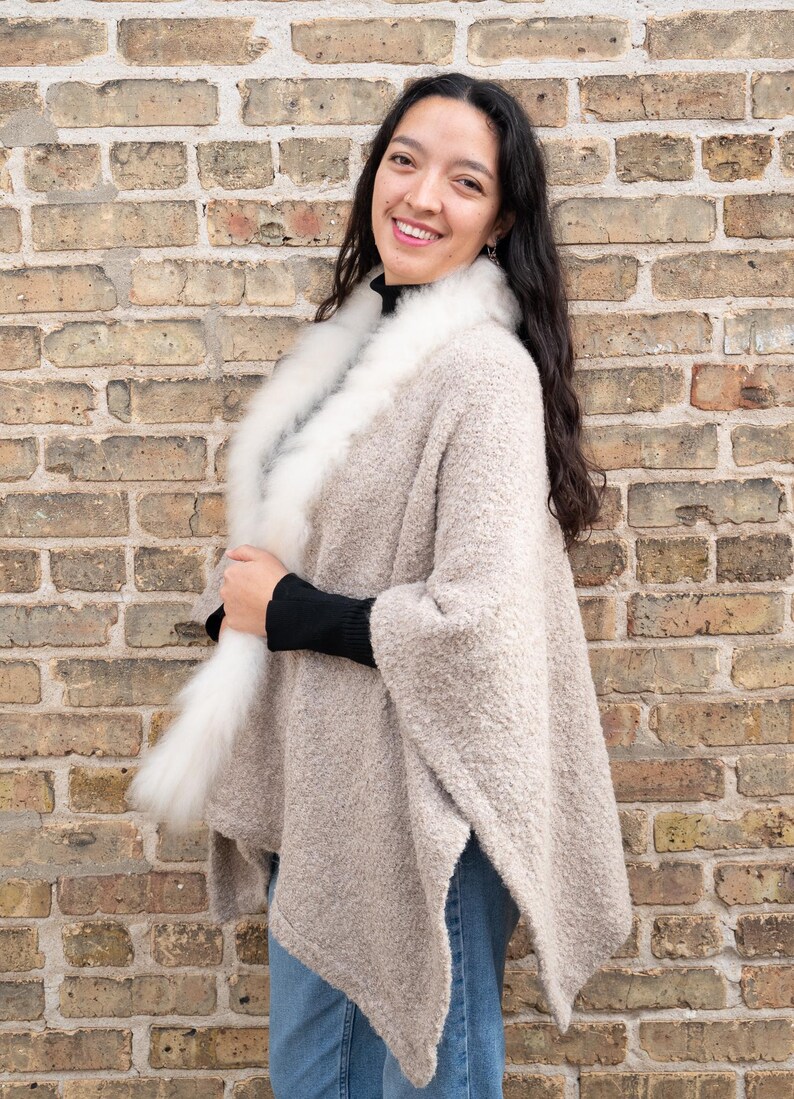 May include: A beige poncho with a white fur collar. The poncho has a textured appearance and is worn over a black top and blue jeans. The background is a brick wall.