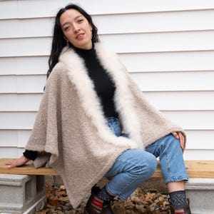 May include: A beige poncho with a white fur trim, worn over a black turtleneck and blue jeans. The poncho has a textured appearance. The person is wearing brown boots and patterned socks. The background is a white wall.