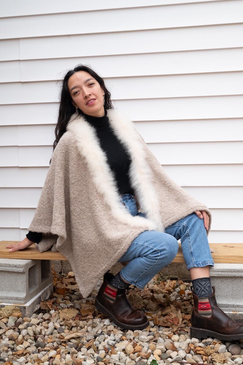 May include: A beige poncho with a white fur trim, worn over a black turtleneck and blue jeans. The poncho has a textured appearance. The person is wearing brown boots and patterned socks. The background is a white wall.