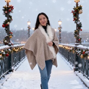 May include: A woman wearing a beige cape with a white fur trim, black turtleneck, and blue jeans walks on a snow-covered bridge. The bridge is decorated with Christmas lights and ornaments. Snowflakes fall in the air.
