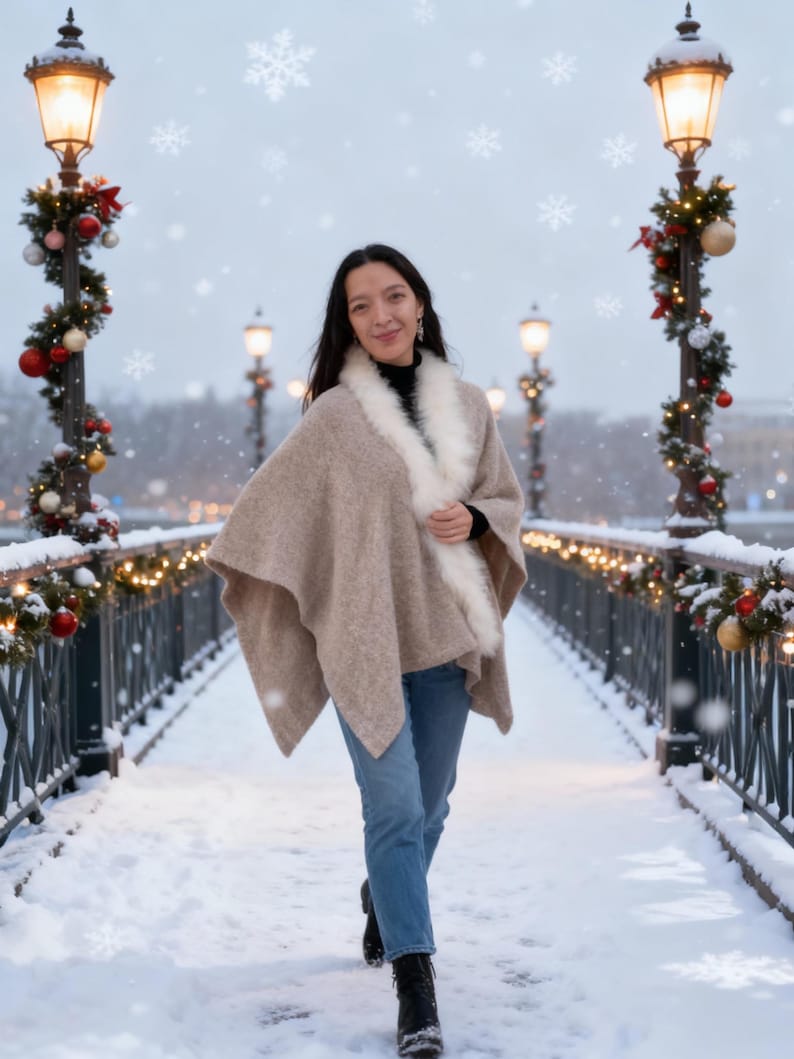 May include: A woman wearing a beige cape with a white fur trim, black turtleneck, and blue jeans walks on a snow-covered bridge. The bridge is decorated with Christmas lights and ornaments. Snowflakes fall in the air.