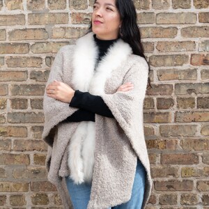 May include: A woman wearing a beige cape with a white fur collar and trim, paired with a black top and blue jeans. The cape has a textured appearance. The background is a brick wall.
