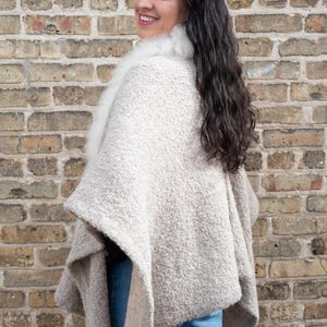 May include: A beige cape with a white fur collar. The cape has a textured, woven appearance and drapes over the shoulders. The person is wearing blue jeans. The background is a brick wall.