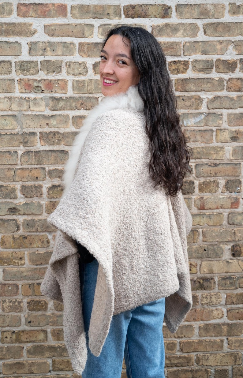 May include: A beige cape with a white fur collar. The cape has a textured, woven appearance and drapes over the shoulders. The person is wearing blue jeans. The background is a brick wall.