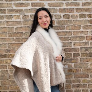 May include: A beige cape with a white fur collar. The cape has a textured, boucle-like appearance. The person is wearing a black top and blue jeans. The background is a brick wall.