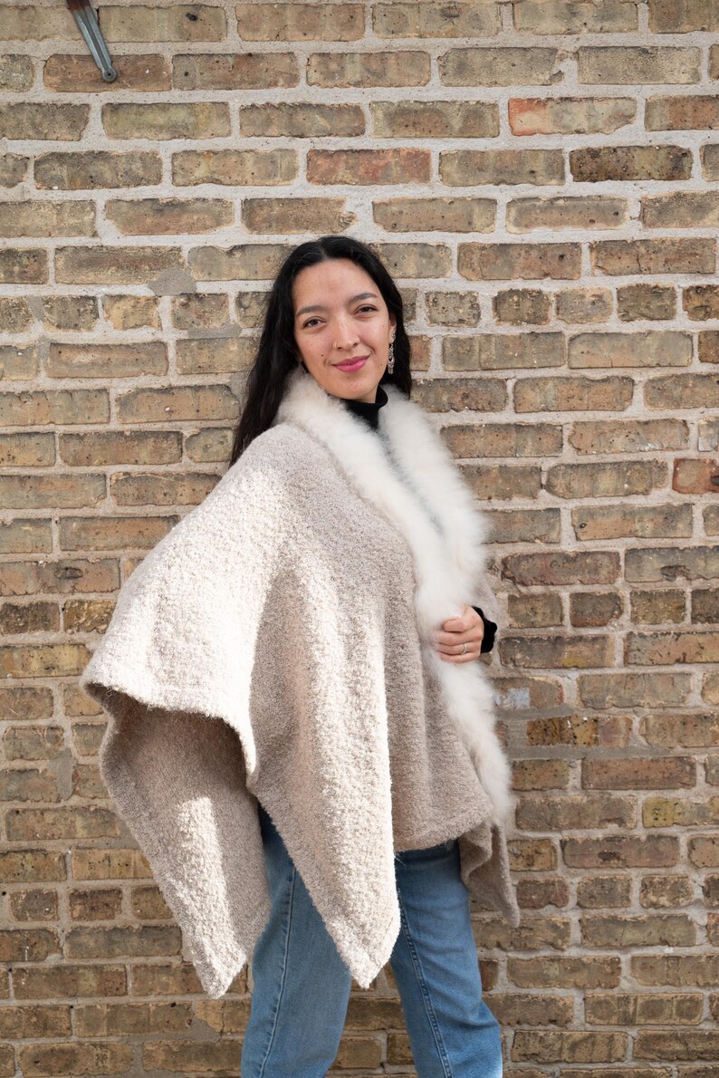 May include: A beige cape with a white fur collar. The cape has a textured, boucle-like appearance. The person is wearing a black top and blue jeans. The background is a brick wall.