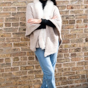 May include: A woman wearing a beige cape with a white fur collar, black turtleneck, and blue jeans. She is standing in front of a brick wall, wearing brown boots.