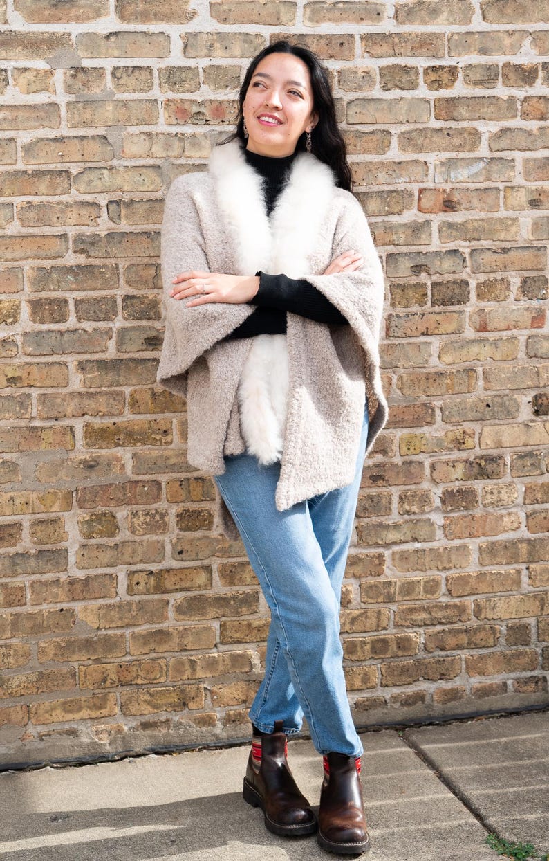 May include: A woman wearing a beige cape with a white fur collar, black turtleneck, and blue jeans. She is standing in front of a brick wall, wearing brown boots.