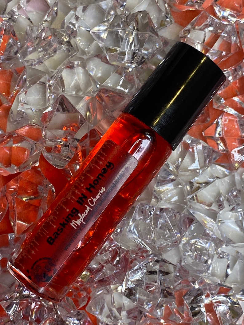 May include: A clear glass roller bottle filled with a reddish-orange liquid, with a black cap. The bottle is labeled "Basking In Honey" and "Moplaed Cherries". The bottle rests on a bed of clear and orange crystal-like shapes.