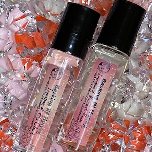 Puede incluir: Dos frascos de perfume roll-on de vidrio transparente con tapas negras. Cada frasco tiene una etiqueta con el texto "Basking In Homes" y "Impression of Baccarat Rouge 540" y "Impression of AG Cloud". Los frascos están rodeados de cristales decorativos transparentes y naranjas.