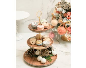 May include: A three-tiered wooden cake stand with a gold metal stand. The stand is decorated with macarons featuring cartoon animal faces and pine cones. The stand is on a white marble surface with a small Christmas tree in the background.