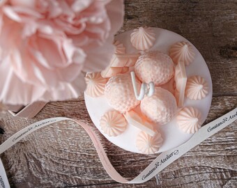 May include: A white ceramic plate with a pink and white floral design candle. The candle is surrounded by a white ribbon with the text "Constance & Atelier" printed on it.
