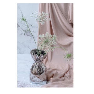 Puede incluir: Un jarr&oacute;n de cer&aacute;mica plateado con forma de bolsa arrugada con un borde rizado, que contiene un ramo de flores blancas.