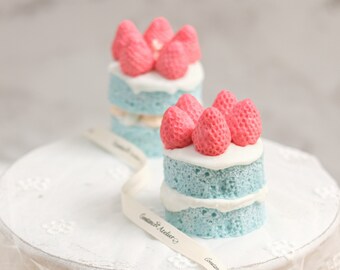 May include: Two miniature cakes decorated with pink frosting and strawberries on a white cake stand with a white lace tablecloth and a white ribbon with the text "Cookies & Keller" printed on it.