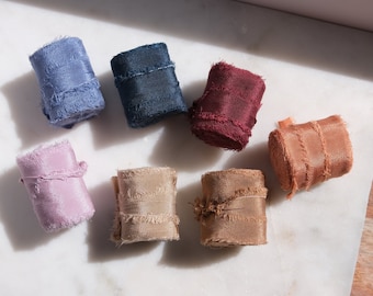 Puede incluir: Siete carretes de cinta con bordes deshilachados en varios colores, incluyendo azul claro, azul oscuro, rojo intenso, morado claro, beige, tostado y cobre. Cada cinta está enrollada en forma cilíndrica, mostrando un acabado texturizado.