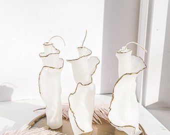May include: Three white candles with gold accents, shaped like abstract organic forms, on a gold tray with a feather boa.