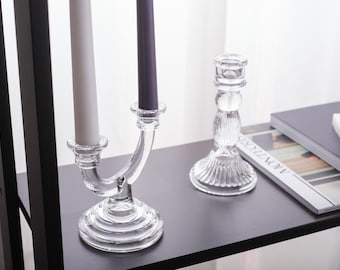May include: Two glass candle holders with candles. The holder on the left has three arms and holds two white candles and one purple candle. The holder on the right is a single arm holder and holds a clear glass candle.