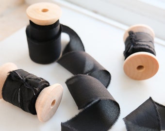 Puede incluir: Varias bobinas de cinta negra de madera están dispuestas sobre una superficie blanca. La cinta se desenrolla de las bobinas, con un borde deshilachado. Las bobinas son de color madera natural y la cinta es de color negro liso.