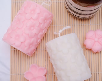 May include: Two decorative candles with a floral design. One candle is pink and the other is white. Both candles are on a bamboo mat.