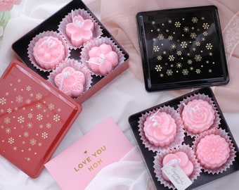 May include: Three pink flower-shaped candles in a black box with a gold snowflake pattern. The box is open and the candles are visible. The candles are in individual white paper cups. The box is on a white surface. There is a pink card with the text "Love You Mom" on the surface.