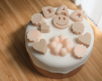 May include: A white cake decorated with pink and beige fondant shapes, including the word "DAD", a smiley face, hearts, and donut-shaped designs.