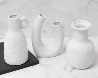 May include: Three white ceramic vases of different shapes and sizes. The vases are on a white marble surface.
