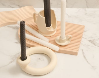 Puede incluir: Un conjunto de tres candelabros de cerámica blanca con velas negras y blancas. Los candelabros tienen diferentes formas, incluyendo una taza, un anillo y un pequeño pedestal. Las velas están todas encendidas y las llamas son visibles.