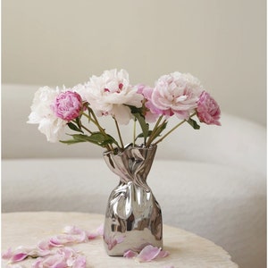 Puede incluir: Un ramo de peon&iacute;as rosas y blancas en un jarr&oacute;n plateado con forma de papel arrugado. El jarr&oacute;n est&aacute; sobre una mesa blanca con p&eacute;talos rosas esparcidos alrededor.