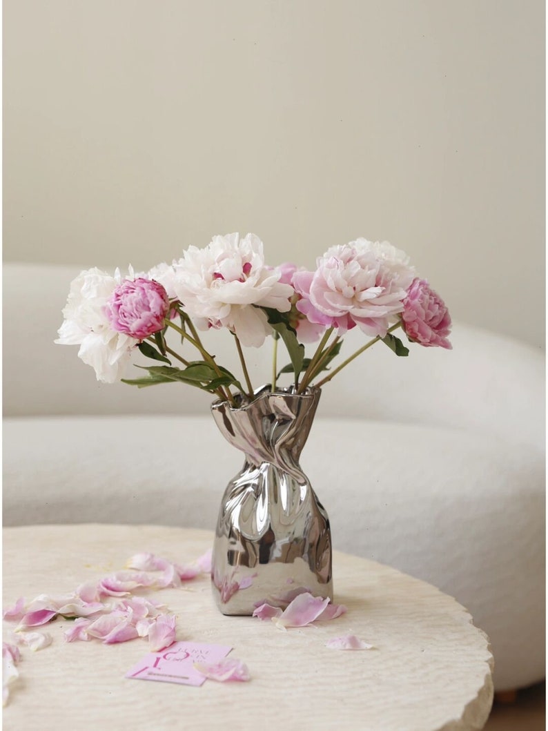 Puede incluir: Un ramo de peon&iacute;as rosas y blancas en un jarr&oacute;n plateado con forma de papel arrugado. El jarr&oacute;n est&aacute; sobre una mesa blanca con p&eacute;talos rosas esparcidos alrededor.