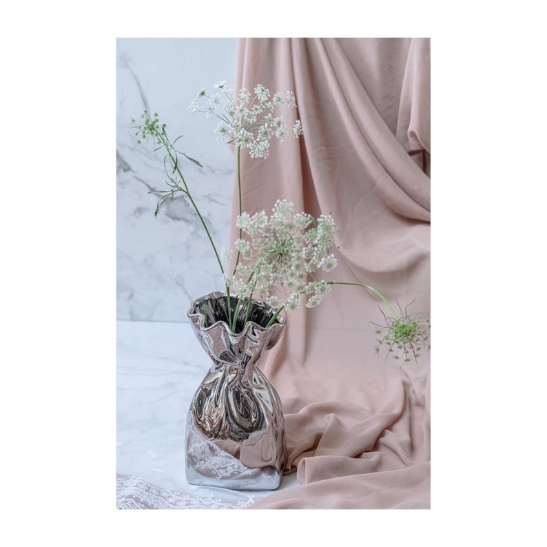Puede incluir: Un jarr&oacute;n de cer&aacute;mica plateado con forma de bolsa arrugada con un borde rizado, que contiene un ramo de flores blancas.