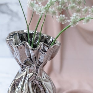 Puede incluir: Un jarr&oacute;n de cer&aacute;mica plateado con un borde rizado y un dise&ntilde;o arrugado y texturizado. El jarr&oacute;n est&aacute; lleno de flores blancas y tallos verdes.
