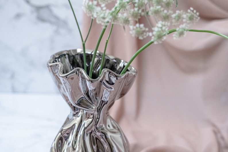 Puede incluir: Un jarr&oacute;n de cer&aacute;mica plateado con un borde rizado y un dise&ntilde;o arrugado y texturizado. El jarr&oacute;n est&aacute; lleno de flores blancas y tallos verdes.