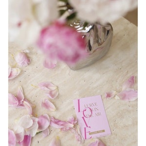 Puede incluir: Tarjeta rosa con las palabras "Love is in the air" y "I love you" en fuente dorada. La tarjeta est&aacute; rodeada de p&eacute;talos de flores rosas.