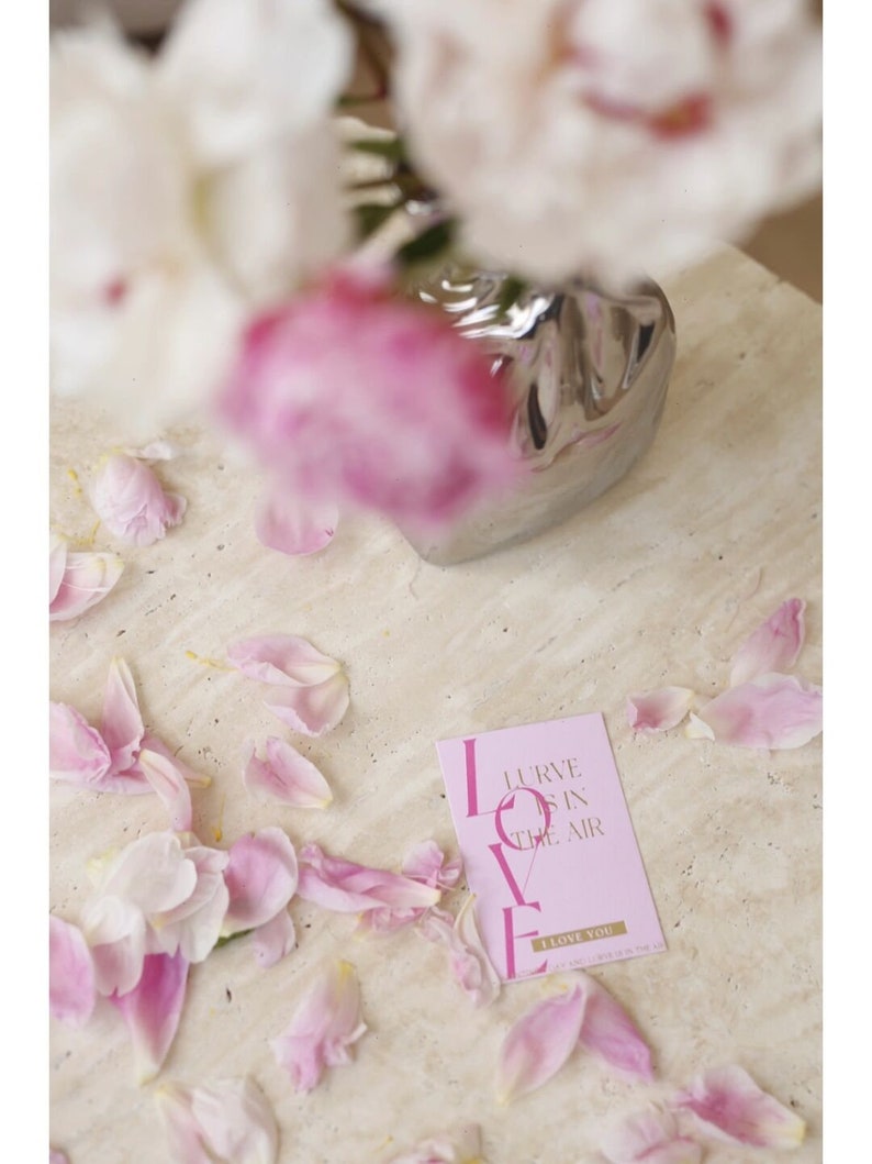Puede incluir: Tarjeta rosa con las palabras "Love is in the air" y "I love you" en fuente dorada. La tarjeta est&aacute; rodeada de p&eacute;talos de flores rosas.