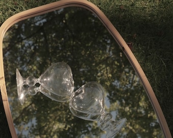 May include: Two clear glass goblets with stems are reflected in a wooden framed mirror. The mirror is reflecting a green grassy area and a tree with green leaves.