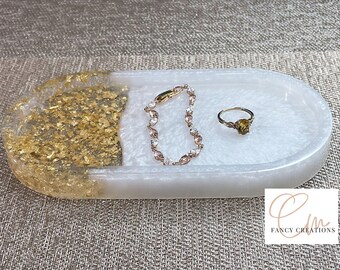May include: White oval resin tray with gold leaf accents. The tray is decorated with a gold ring and a silver bracelet. The tray is on a brown textured surface. "Fancy Creations" is written in the bottom right corner of the image.