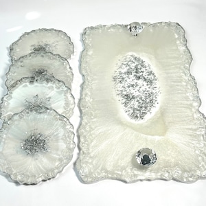 May include: A set of white resin coasters and a serving tray. The coasters and tray have a silver glitter design in the center and around the edges. The tray has two clear crystal knobs.