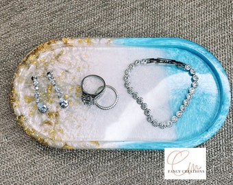 May include: A white and blue oval resin tray with gold flecks. The tray is filled with jewelry, including a bracelet, earrings, and two rings.