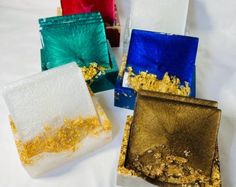 May include: Set of six coasters in various colors, including white, green, blue, and brown. Each coaster has gold leaf accents and is displayed in a matching holder.