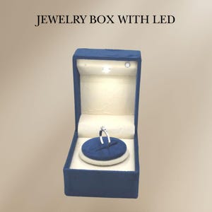 May include: A blue velvet jewelry box with a white LED light inside. The box has a white velvet interior and a blue velvet ring holder. A silver ring with a small diamond is displayed in the ring holder.