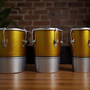 Könnte beinhalten: Drei gold-silberne Conga-Trommeln in einer Reihe. Die Trommeln haben einen metallisch goldenen oberen Teil und einen silbernen unteren Teil, mit silberfarbener Hardware. Sie stehen vor einer Backsteinmauer und auf einer Holzoberfläche.